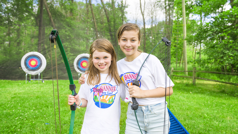 Two girl campers with bows and arrows smiling by the archery field