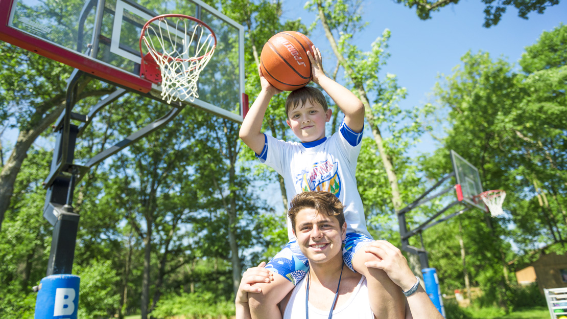 Boy camper holding a basketball up to the hoop while sitting on male staff members shoulders