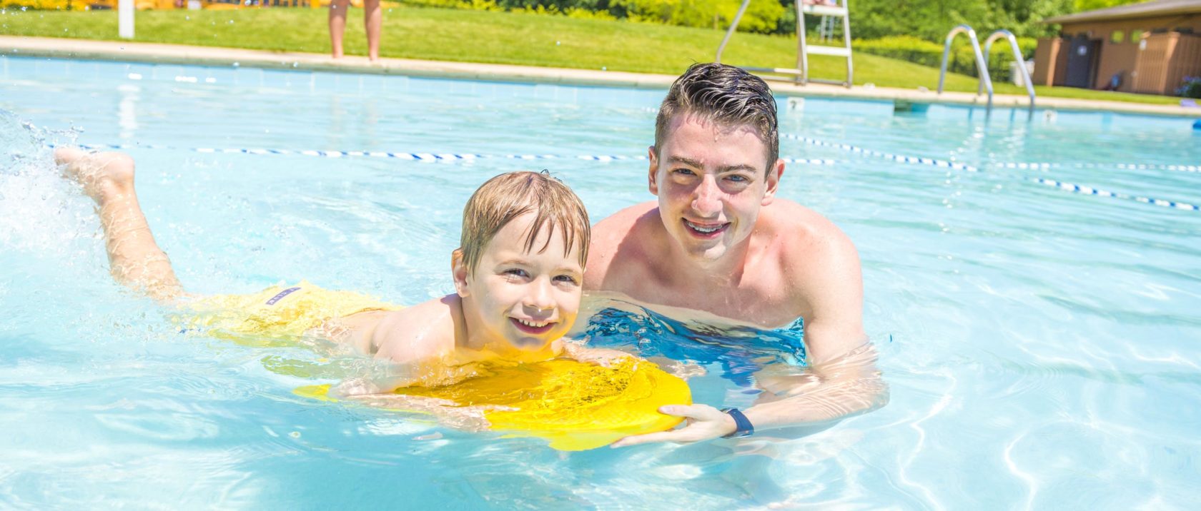 Male staff member teaching a boy camper how to swim on a boogie board in the pool