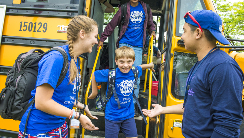 Boy camper walking off of the bus with counselors greeting him