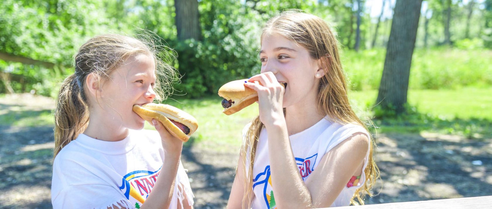 Two girl campers eating hot dogs at a table