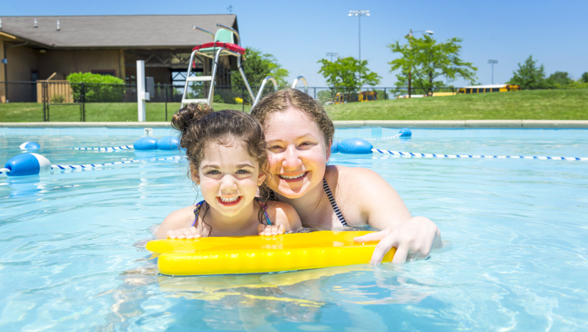 Female staff member teaching girl camper how to swim in the pool with a boogie board