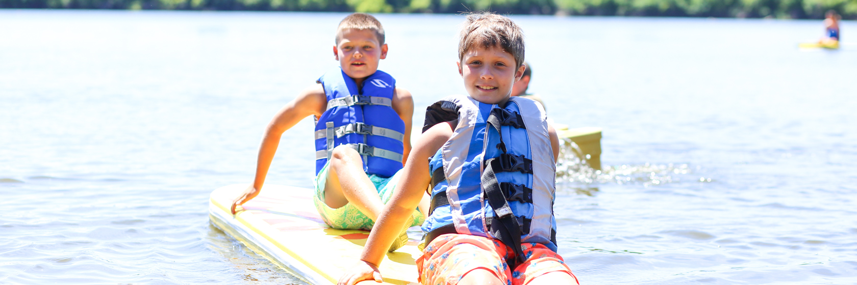 Two boy campers sitting on a paddle board in the lake