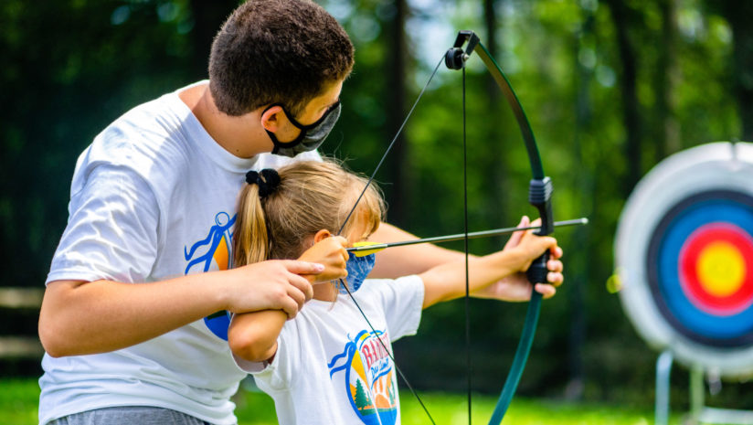 Male counselor leads camper at archery with masks on